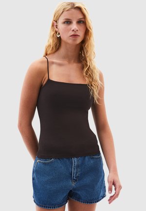 Giovane donna con capelli biondi lunghi che indossa un top nero con spalline sottili e pantaloncini di jeans blu, su uno sfondo grigio neutro.
