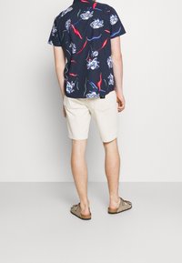 Camisa de manga corta azul marino con un patrón floral en blanco y rojo, combinada con pantalones cortos beige claros y sandalias marrones.
