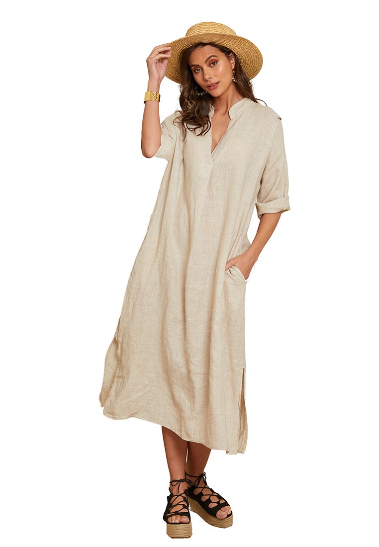 Leinenkleid in hellbeige mit V-Ausschnitt, aufgerollten Ärmeln, seitlichen Taschen und Schlitzen am Saum, kombiniert mit schwarzen, geflochtenen Espadrilles-Sandalen.