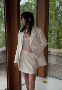 Ensemble composé d'un blazer blanc texturé et d'une mini jupes à motif floral, associé à un haut transparent. La tenue présente une silhouette ajustée et des manches larges.