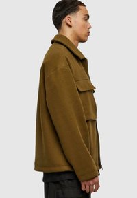 Veste en polaire marron avec un col haut, fermeture éclair à l'avant et une poche poitrine bien visible. Elle présente une coupe décontractée et des manches longues.