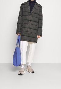 Personne portant un manteau boutonné rayé gris et vert, un pantalon blanc court, des baskets beiges, et tenant un sac fourre-tout bleu à la main.