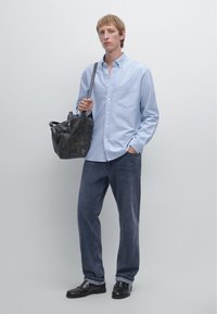 Light blue button-up shirt, dark blue jeans, black shoes, holding a black leather bag. Simple, classic design with a front pocket on the shirt.