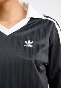 Μαύρη αθλητική μπλούζα με μακριά μανίκια και κάθετες ρίγες, λευκό γιακά και το λογότυπο της Adidas κεντημένο σε λευκό στο στήθος.