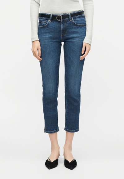 Femme portant un jean slim bleu court avec une ceinture noire et des chaussures slingback noires à bout pointu, debout devant un fond blanc uni.