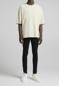 Camiseta oversize de algodón color beige claro con manga corta, combinada con jeans pitillo negros y zapatillas blancas con un detalle en el talón en color contrastante.