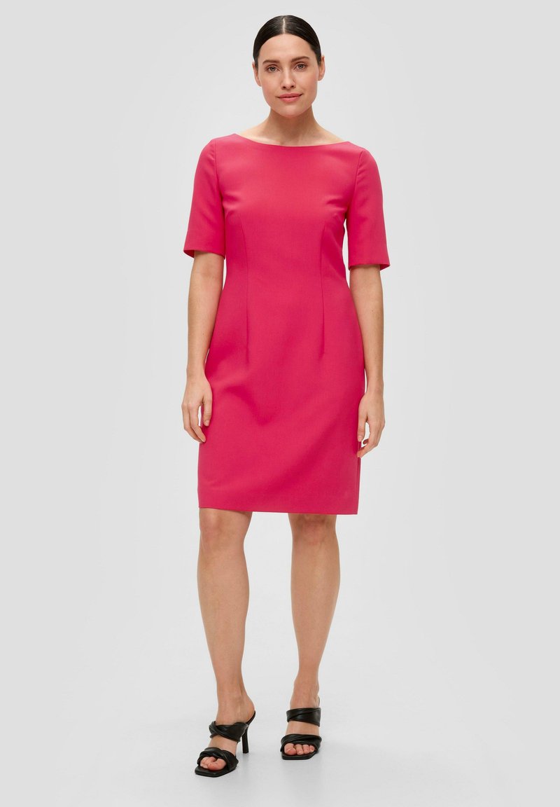 Robe rose ajustée avec des manches courtes, un col rond et des plis subtils. Portée avec des sandales à talons noirs sur un fond neutre.