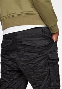 Personne portant un pantalon cargo noir avec plusieurs poches et un sweat-shirt vert, main dans la poche, vue de la taille vers le bas.
