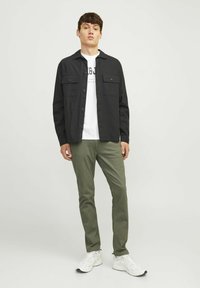 Camicia nera con bottoni e due tasche sul petto, indossata sopra una maglietta bianca con grafica, abbinata a pantaloni verde oliva e sneakers bianche.
