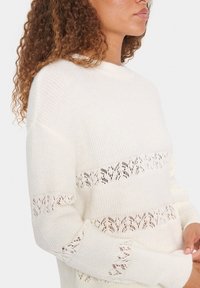 Cream-colored sweater with long sleeves, featuring a ribbed texture and lace patterns across the body and sleeves. Round neckline.