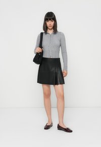 Siva zip-up pulover s rebrastim vzorcem, črna plisirana krilo iz umetnega usnja in burgundske lakirane balerinke. Model drži majhno črno torbico.