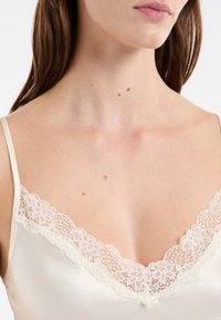 Cou de femme et partie supérieure de la poitrine avec une peau claire, portant un caraco en satin ivoire avec des dentelles et des grains de beauté visibles sur la peau.