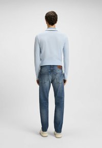 Polo a maniche lunghe di colore azzurro abbinato a jeans blu scuro. Dettagli notevoli includono un patch con logo sulla cintura e un tessuto strutturato.
