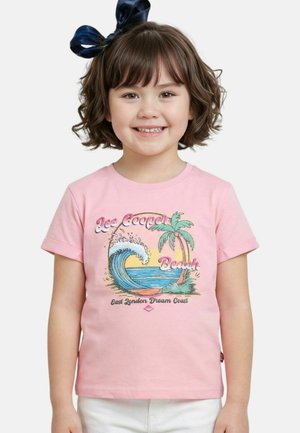 Jeune fille souriante aux cheveux bruns courts avec un nœud marine, portant un t-shirt rose avec un motif de vague océanique, un palmier et un coucher de soleil, ainsi qu'un pantalon blanc.