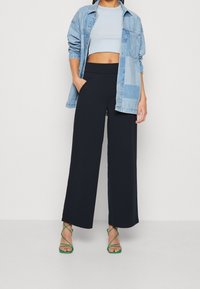 Džínová košile přes světle modré crop top, doplněná širokými černými kalhotami a zelenými sandály na řemínky. Má ležérní střih a vrstvený vzhled.