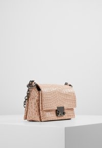 Piccola borsa a tracolla beige con texture a coccodrillo, tracolla in catena argentata e chiusura in metallo su un piedistallo bianco contro uno sfondo neutro.