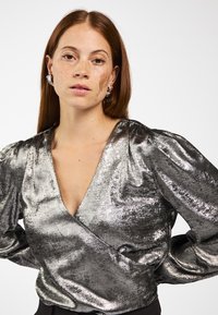Vrouw met lang rood haar en sproeten draagt een zilverkleurige metallic blouse met pofmouwen en zilveren ringoorbellen, handen op de heupen.
