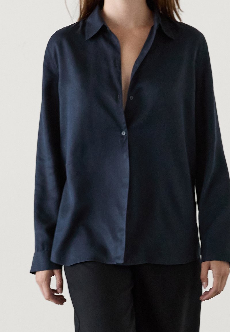 Camicia in satin blu navy con bottoni, colletto classico, maniche lunghe e vestibilità rilassata. Presenta una leggera lucentezza e un design minimale.