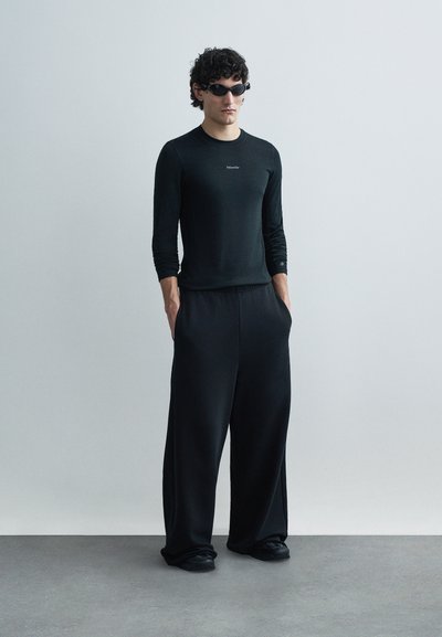 Čierna košeľa s dlhými rukávmi a nohavice so širokými nohavicami, oboje vyrobené z hladkej, ľahkej látky. Doplnky zahŕňajú tmavé slnečné okuliare a čierne topánky.