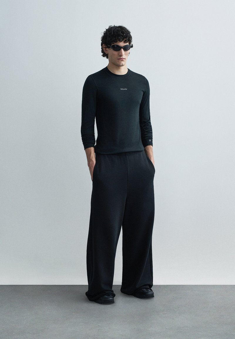 Schwarzes Langarmhemd und weit geschnittene Hose, beide aus glattem, leichtem Stoff. Accessoires sind dunkle Sonnenbrillen und schwarze Schuhe.