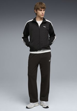 Ung man som bär svartvit Puma-träningsdräkt och vita sneakers, står med händerna i fickorna och tittar åt sidan mot en grå bakgrund.