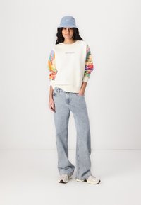 Sudadera con mangas largas coloridas y estampadas, cuerpo blanco y logo "RIP CURL"; combinada con jeans de pierna ancha de color azul claro y un sombrero tipo bucket azul.