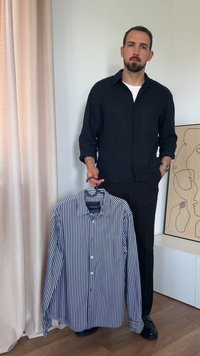 Koszula zapinana na guziki w granatowo-białe pionowe paski, z kołnierzykiem i długimi rękawami, trzymana przez osobę w czarnej koszuli zapinanej na guziki.