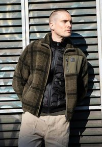 Olijfgroene geruite fleecejack met een zwarte geïsoleerde voorkant en een ritssluiting op de borst, gecombineerd met lichtbeige gestructureerde broek.