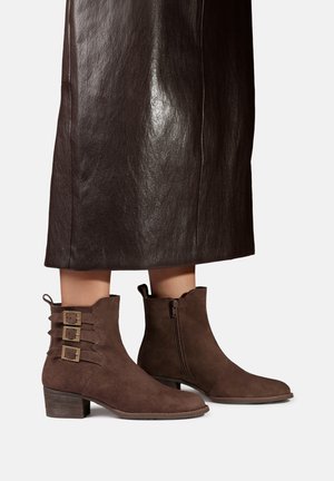Bottines en daim marron avec trois boucles latérales et talons blocs bas portées avec une jupe en cuir marron foncé.