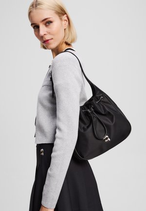 Sac seau en nylon noir avec fermeture à cordon, présentant une finition lisse et un détail de logo. Le modèle porte un pull gris et une jupe noire.