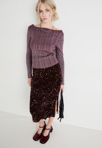 Haut à manches longues côtelé bourgogne associé à une jupe midi brune ornée de sequins avec une fente sur le côté. Le modèle porte un sac noir.