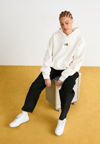 Κρεμ φούτερ με μικρό μαύρο λογότυπο, συνδυασμένο με μαύρο παντελόνι και λευκά sneakers. Μοντέλο καθισμένο σε μεταλλικό κουτί πάνω σε κίτρινο δάπεδο.