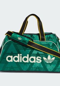 Zöld sporttáska geometriai mintával, fehér "adidas" logóval, állítható fekete pánttal sárga részletekkel. Tartós anyagból készült.