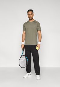 Szara sportowa koszulka, czarne spodnie dresowe, białe trampki. Trzyma rakietę tenisową i żółtą piłkę tenisową. Gładka tkanina, luźny krój.