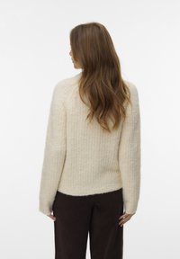 Frau mit langem braunem Haar, die einen cremefarbenen, strukturierten Pullover und dunkelbraune Hosen trägt, mit dem Rücken zur Kamera vor einem schlichten weißen Hintergrund.