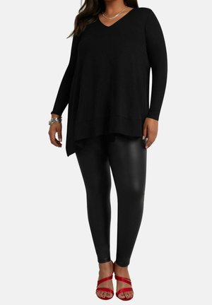 Žena oblečená v dlouhém černém topu s výstřihem do V, černých legínách s koženým vzhledem, stříbrných náramcích a červených otevřených sandálech na podpatku s pásky.