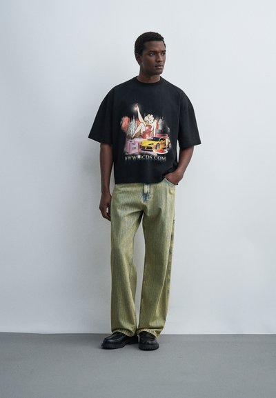 Άνδρας που φοράει μαύρο φαρδύ γραφικό μπλουζάκι με σχέδιο καρτούν και αυτοκινήτου, φαρδιά ανοιχτοπράσινα τζιν και μαύρα παπούτσια, στέκεται μπροστά σε απλό τοίχο.