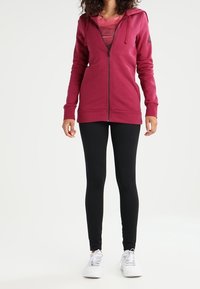 Burgundská zipová mikina s kapucí, kombinovaná s černými legínami a bílými teniskami. Mikina má hladkou texturu a přední kapsy.