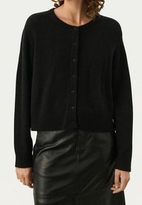 Svart cardigan i mjukt material, med rund halsringning, långa ärmar och knäppning fram. Bärs över en lädrokjol.