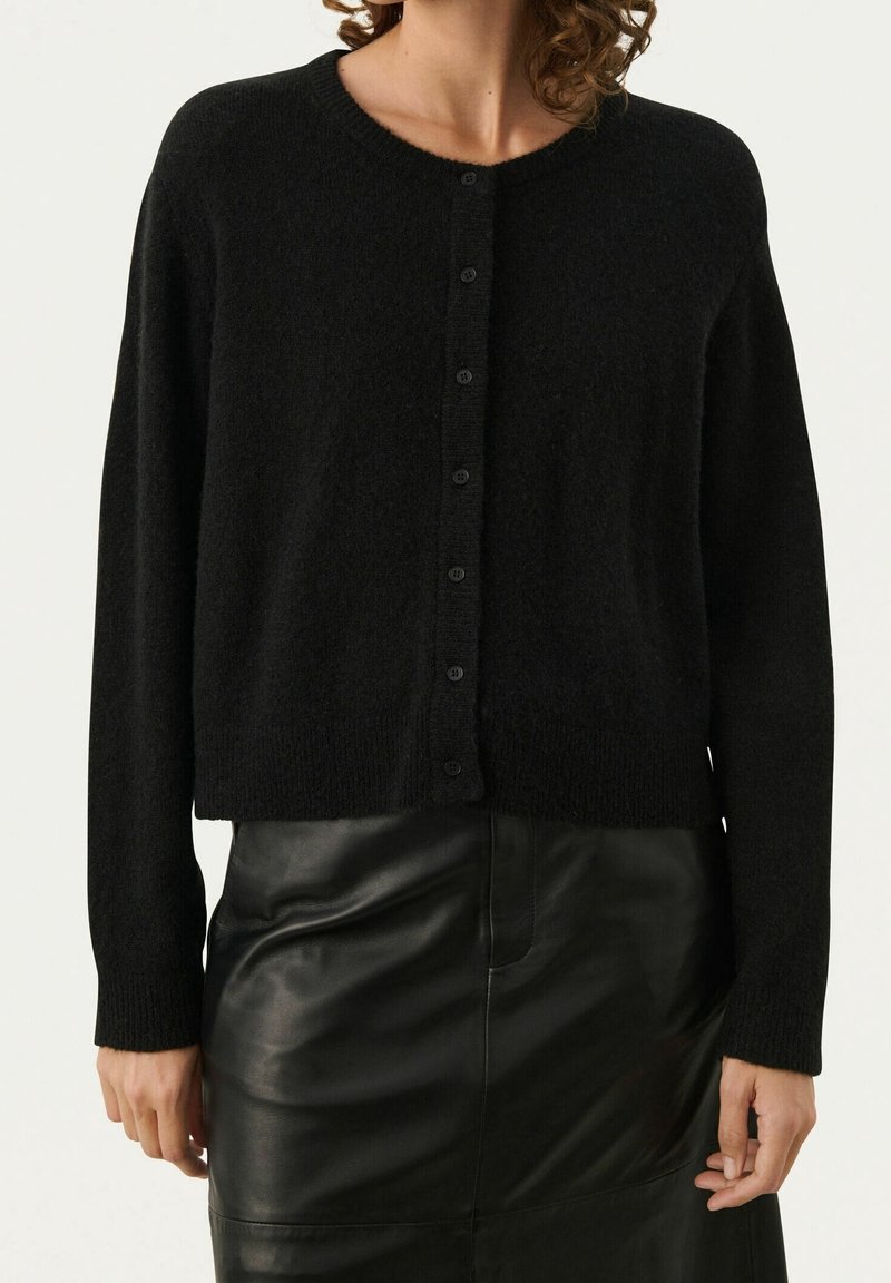 Svart cardigan i mjukt material, med rund halsringning, långa ärmar och knäppning fram. Bärs över en lädrokjol.