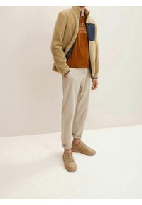 Béžová fleecová bunda na zip s námořnickou kapsou, hnědé tričko, světle béžové kalhoty a světle hnědé tenisky. Texturovaná tkanina; volný střih.