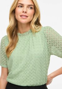 Blouse vert clair présentant un motif texturé, manches courtes et encolure à volants. Tissu doux avec une coupe décontractée.