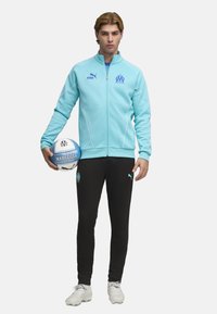 Chaqueta deportiva azul claro con cremallera, detalles y logo en blanco, pantalones negros, sujetando un balón de fútbol azul. Presenta el logo en los pantalones y el balón.