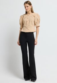 Beige top med puffærmer og seks knapper, parret med sorte tætsiddende bukser. Toppen har en tekstureret stof og et cropped design.