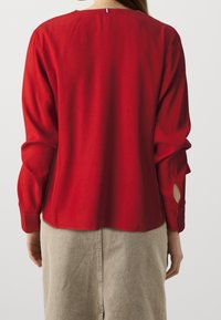 Blusa roja de manga larga con cuello redondo y puños abotonados, que presenta detalles de corte en las mangas. Tejido suave, con un ajuste ligeramente holgado.