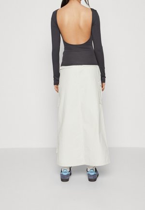 Osoba ubrana w czarną bluzkę z długim rękawem z głębokim odsłoniętym plecami, białą długą spódnicę z kieszeniami, białe skarpetki i niebiesko-szare trampki, widziana od tyłu.