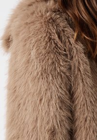 Manteau en fausse fourrure beige et duveteux avec une texture douce visible, porté par une personne aux longs cheveux bruns sur un fond clair.