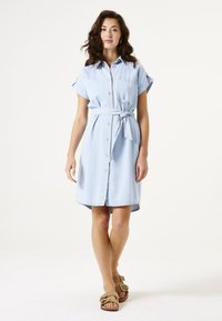 Hellblaues Denim-Kleid mit kurzen, umgeschlagenen Ärmeln, einer Fronttasche, Knopfverschluss und einem gebundenen Taillengürtel. Wird mit braunen Sandalen getragen.