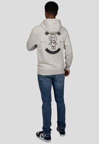 Felpa beige con una grafica di una bambola cucita e testo sul retro. Abbinata a jeans blu e sneakers nere con dettagli bianchi.