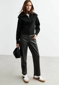 Svart texturerad stickad tröja med dragkedja i kragen, tillsammans med gråa mockaläderbyxor. Vita och svarta sneakers, håller en svart handväska.